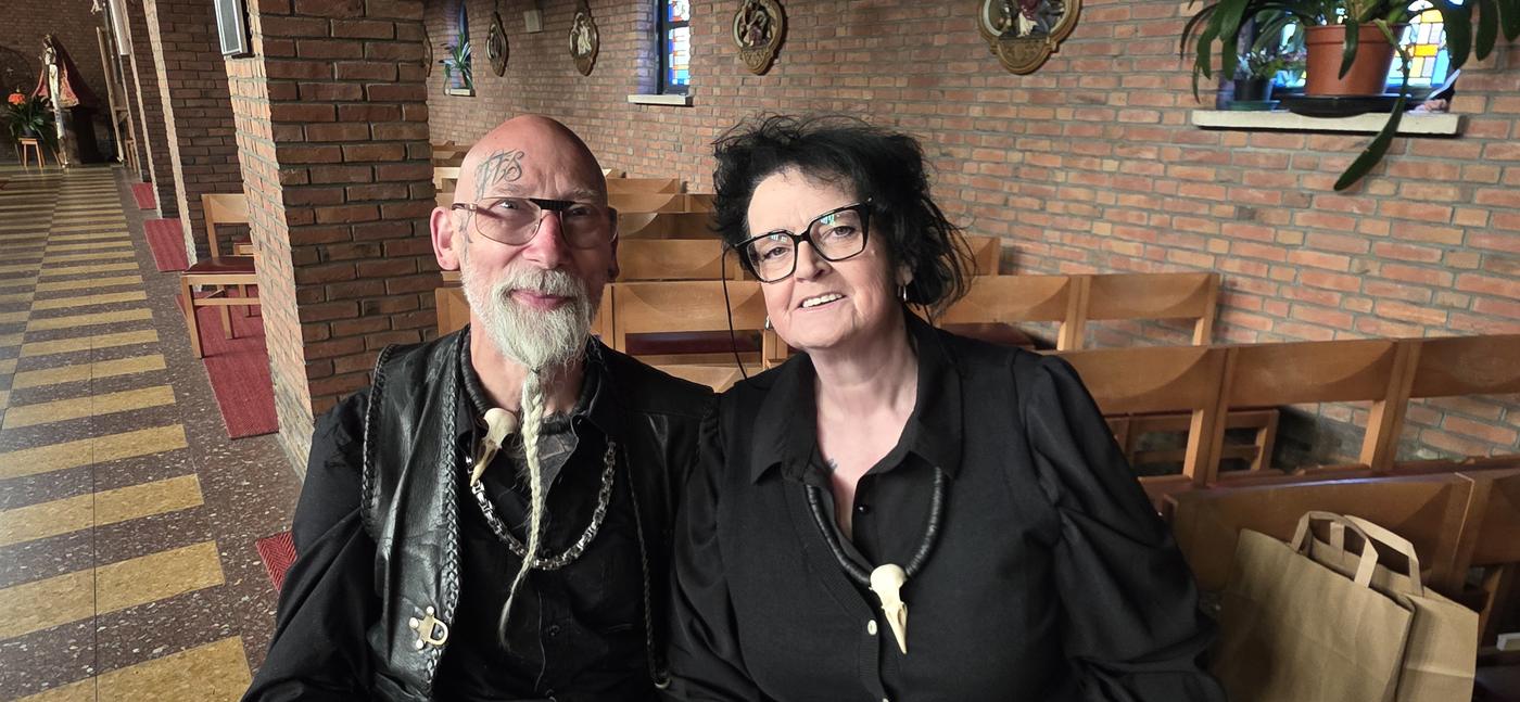 Elly and Flipke seated together in a warm lit church hall, both dressed in black.